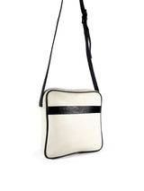 Square medium cross body bag. Cream colored leather with black leather accent stripe on front panel and black piping. Lightweight black cotton lining. Silver buckle on adjustable black leather cross body strap. Slip pocket on back panel. Upcycled, circular, sustainable. 3/4 view.