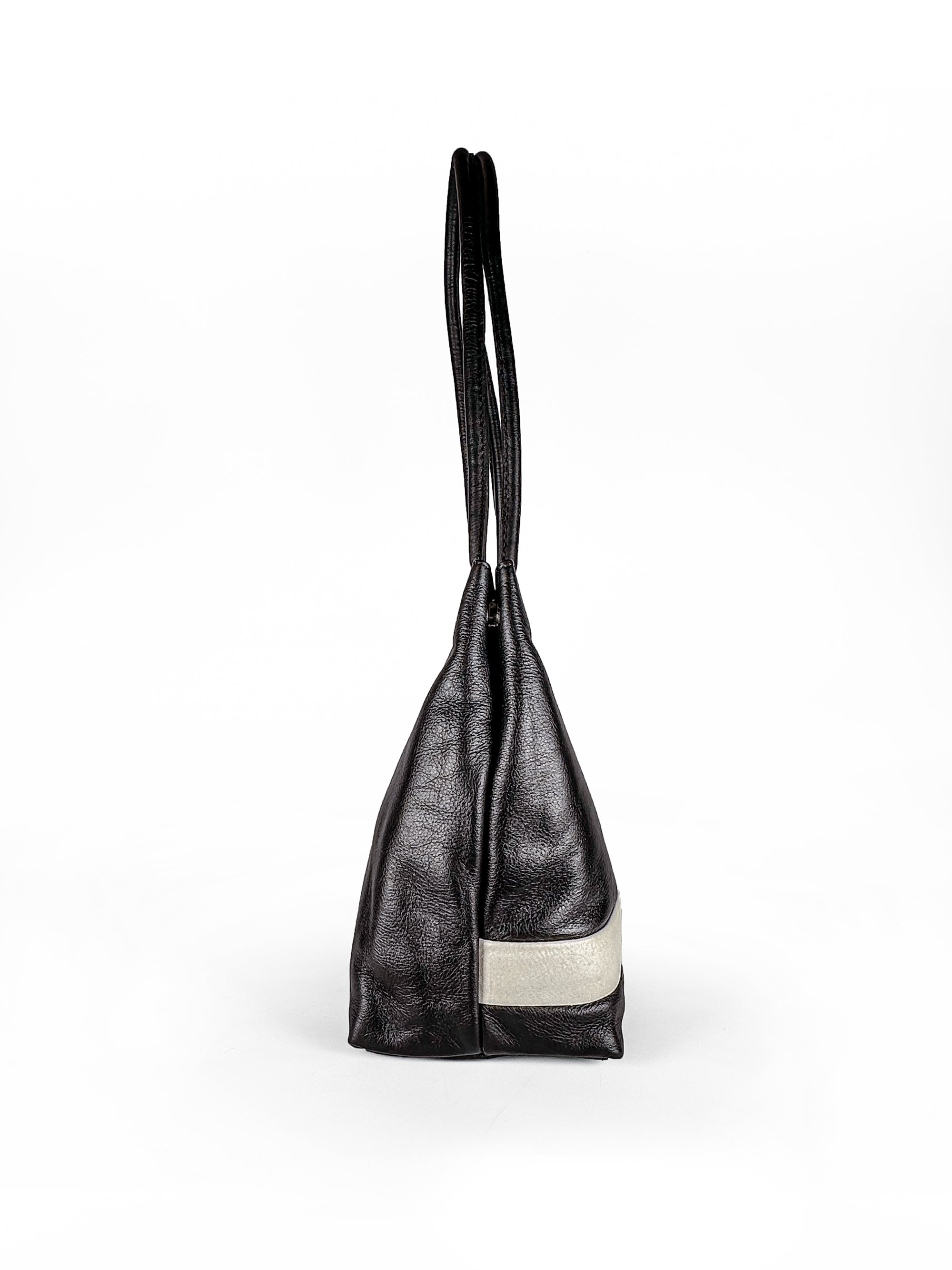 Black leather medium sized handbag, with tapered top. Inlaid white leather accent stripe on front panel. Magnet depth expansion. Chic padded pipe shoulder straps. Upcycled, circular, sustainable. Side view.